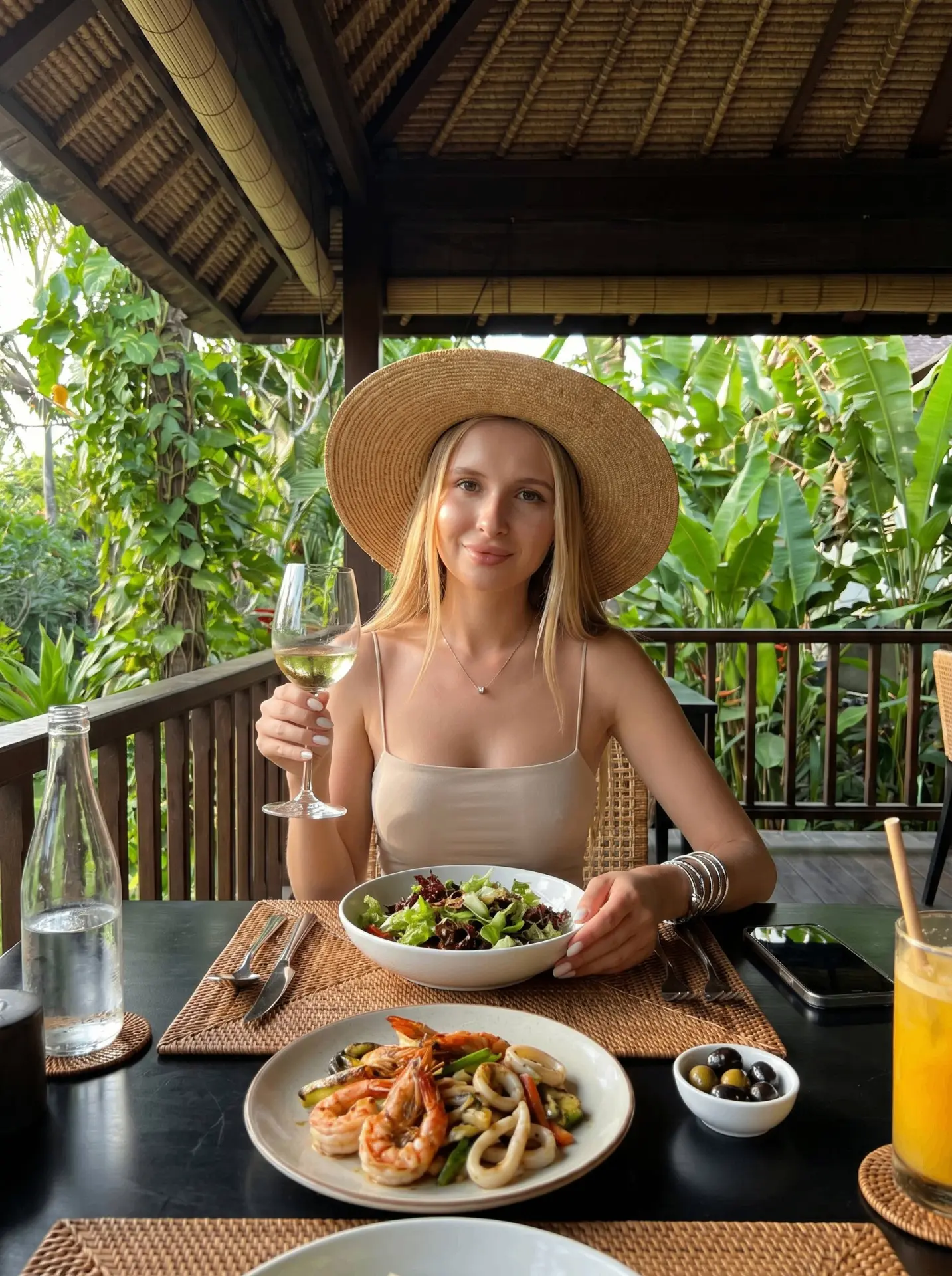 A straight-on, medium shot captures a woman wearing a large, woven straw hat and a light beige spagh
