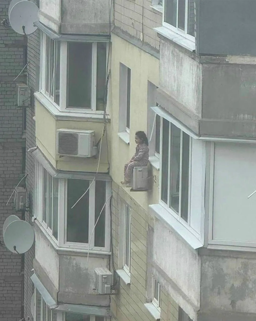 На этом фото девочка, сидящая на кондиционере за окном.