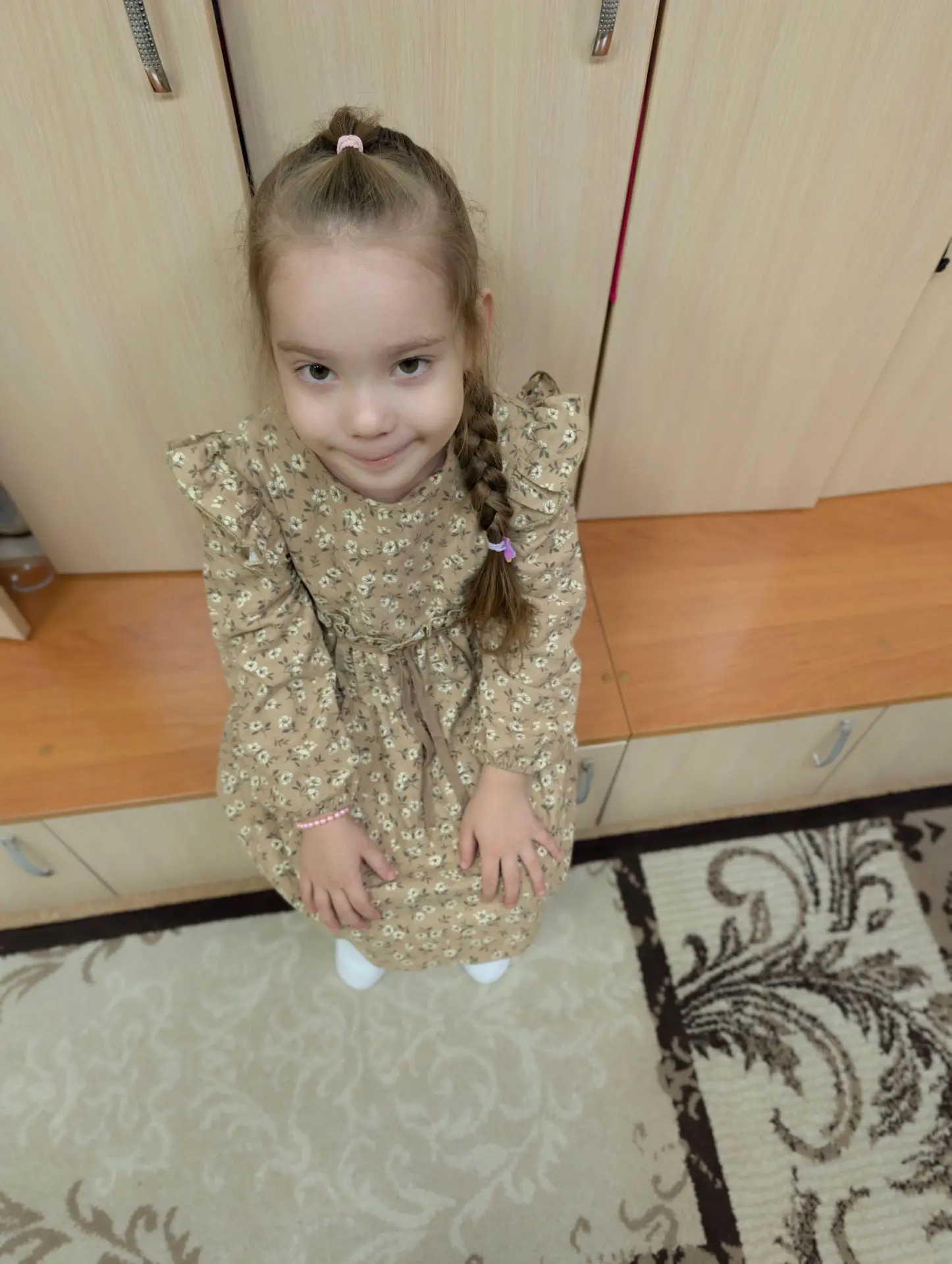 На фотографии изображена **девочка** дошкольного возраста. Она стоит в помещении, скоре...